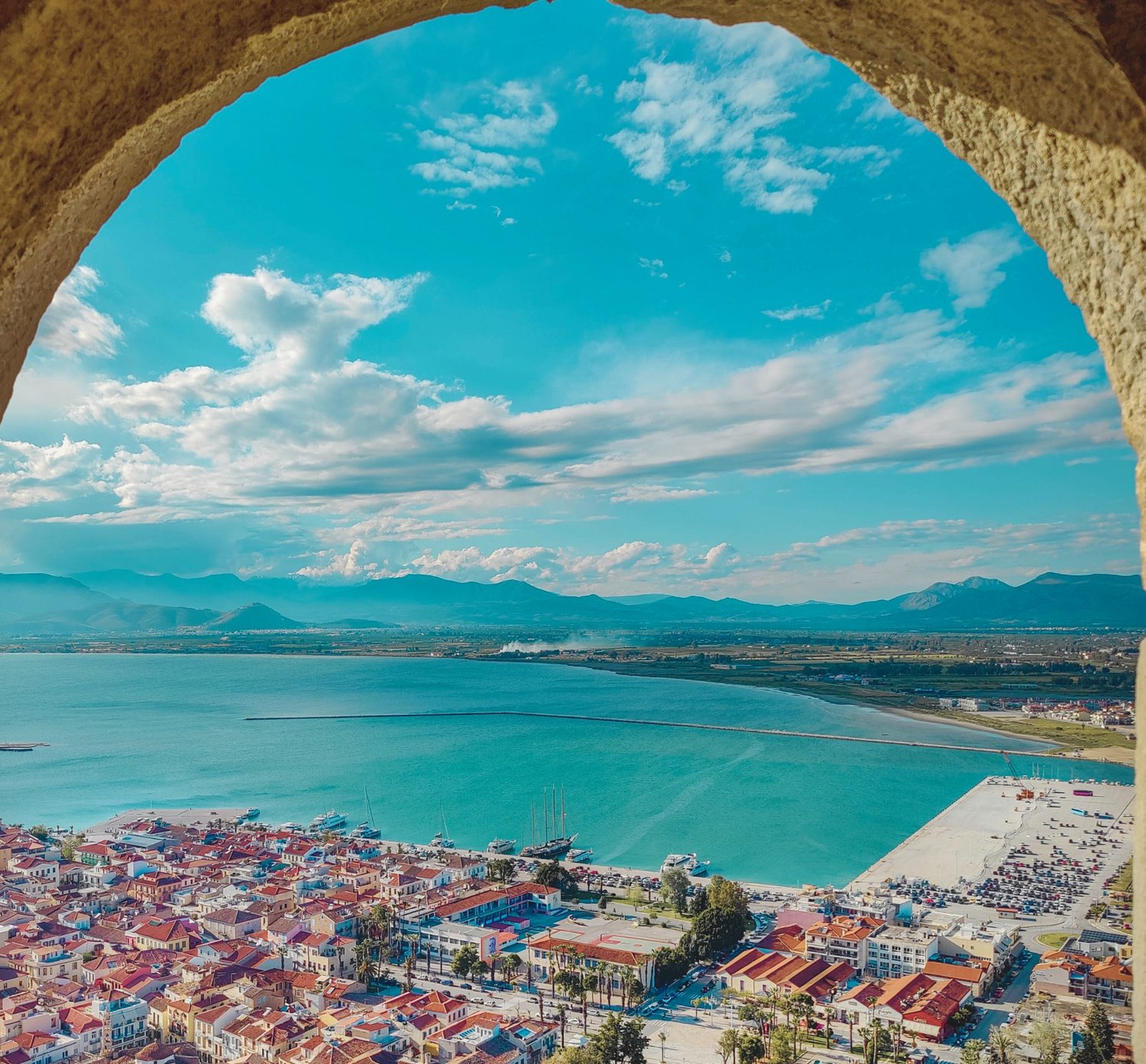 Coastal town and harbor framed by a stone arch, with mountains beyond